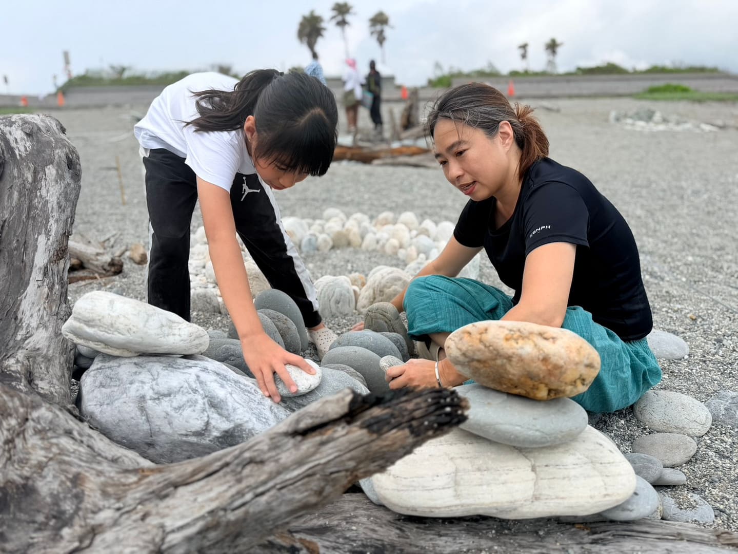Mother and daughter kneeling on a pebbled beach, stacking smooth stones into a small rock formation.