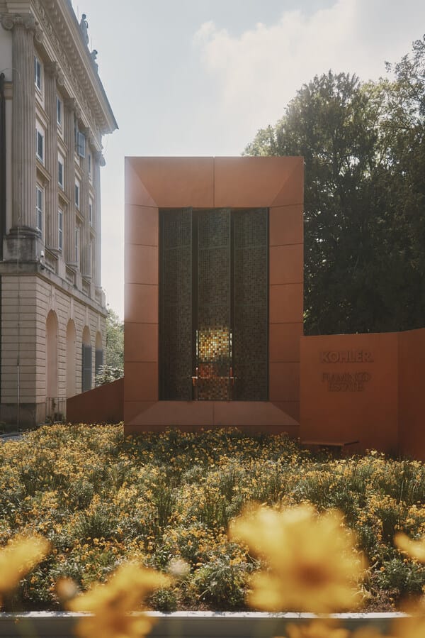 Orange rectangular monument with a metal lattice center, set among yellow flowers; a historic building looms to the left and trees are in the background.