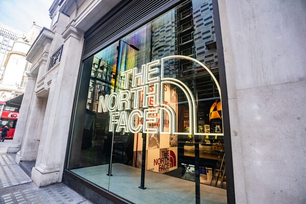 The North Face storefront with a large neon logo in the window on a city sidewalk outside.
