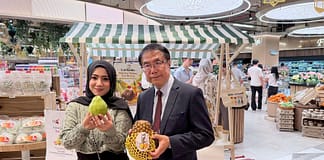 台南甜征服大馬!黃偉哲赴吉隆坡推廣紅心芭樂、冬瓜茶,鳳梨首波秒殺 Two people pose for a photo inside a bright supermarket, a woman in a headscarf holding a large green fruit and a man in a suit holding a pineapple wrapped in protective netting, with a striped market stall and produce in the background.