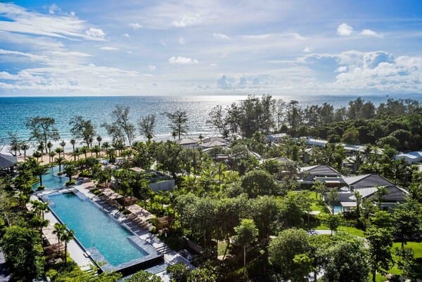 Aerial view of a tropical resort with a long pool, palm trees, and white buildings beside a blue sea on a sunny day.
