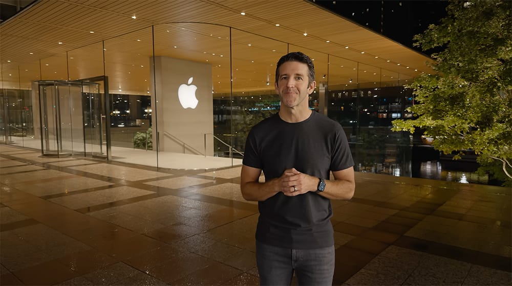 Man in a black t-shirt and jeans stands outside an Apple Store at night, hands clasped in front.