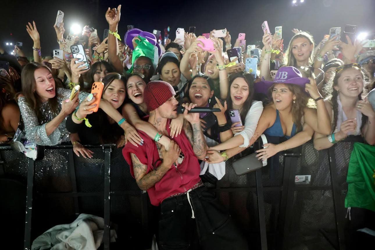 Male singer in a red beanie performs at a crowded concert, fans reach over the barrier with phones raised around him.