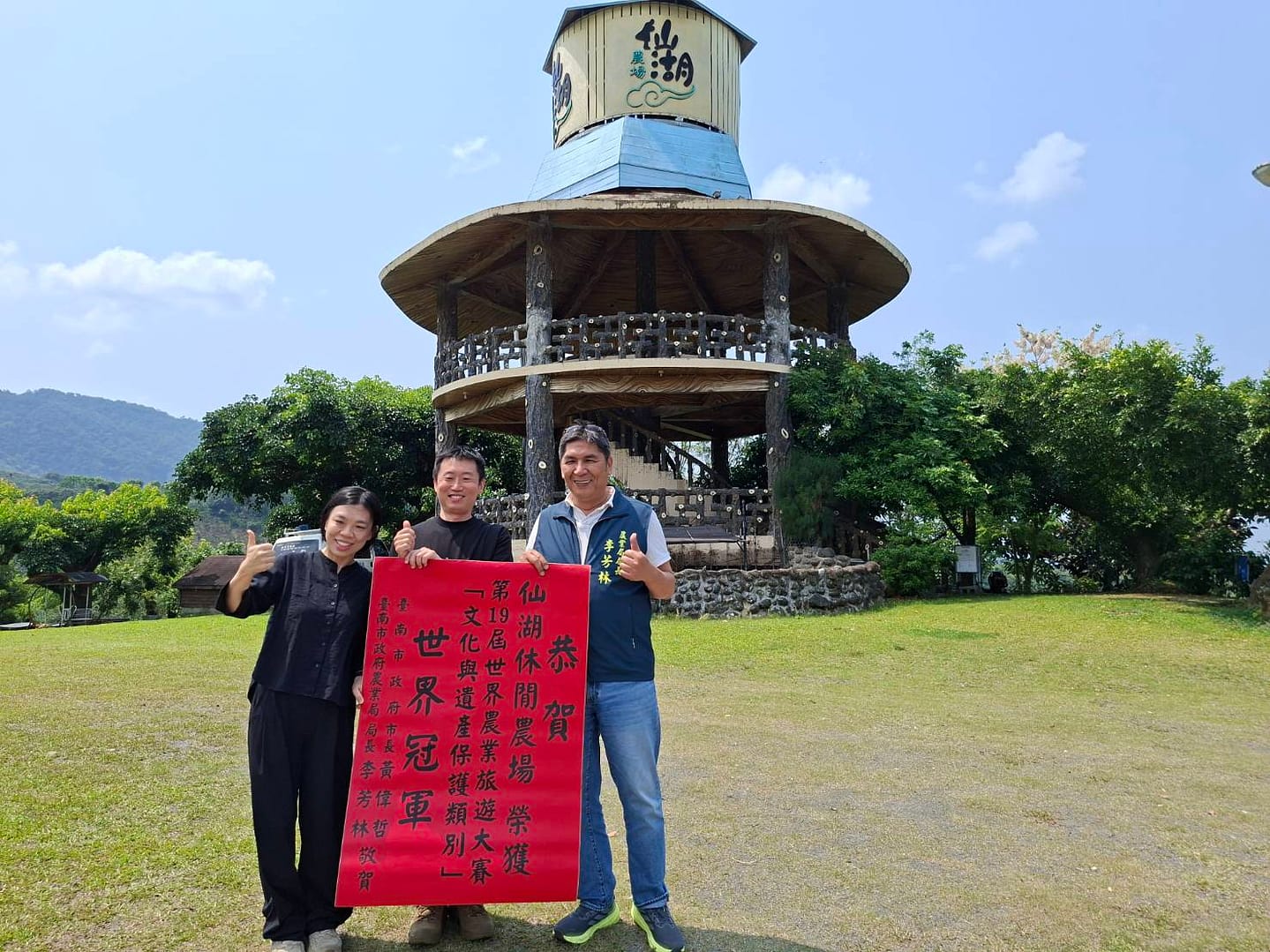 Three people pose on a grassy field, holding a red banner with Chinese characters, in front of a circular wooden pavilion under a blue sky.