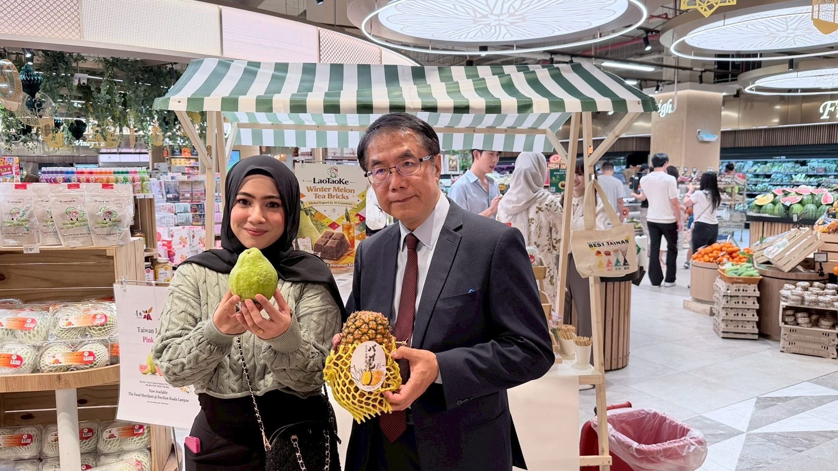 Two people pose for a photo inside a bright supermarket, a woman in a headscarf holding a large green fruit and a man in a suit holding a pineapple wrapped in protective netting, with a striped market stall and produce in the background.