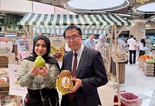 台南甜征服大馬!黃偉哲赴吉隆坡推廣紅心芭樂、冬瓜茶,鳳梨首波秒殺 Two people pose for a photo inside a bright supermarket, a woman in a headscarf holding a large green fruit and a man in a suit holding a pineapple wrapped in protective netting, with a striped market stall and produce in the background.