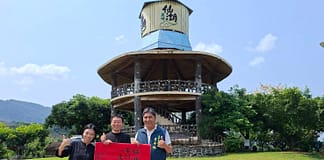 臺南之光!仙湖休閒農場勇奪第19屆世界農業 旅遊大賽「文化與遺產保護類別」世界冠軍 Three people pose on a grassy field, holding a red banner with Chinese characters, in front of a circular wooden pavilion under a blue sky.