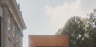 Kohler 與 Flamingo Estate 於米蘭設計週打造融合儀式感、自然與工藝的雕塑式浴場 Orange rectangular monument with a metal lattice center, set among yellow flowers; a historic building looms to the left and trees are in the background.